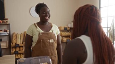 İki siyahi kadın fırında dostça bir gülümsemeyle etkileşimde bulunuyorlar. Kapalı bir dükkanda sıcak bir atmosfer yaratıyorlar.