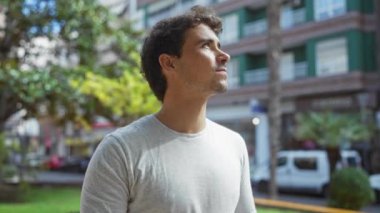Şehir parkında yeşil yeşillik ve şehir arkaplanı olan sıradan giyinmiş genç bir adam, güneşli bir ortamın keyfini çıkarırken düşünceli bir ifadeyle yukarı bakıyor.