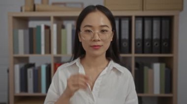 Ofisinde rafları ve klasörleri olan gözlük takan bir kadın beyaz gömlek giyerken profesyonel ve odaklanmış bir görünüm sergiliyor.