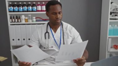 Beyaz önlüklü genç adam klinikte oturmuş arka planda ilaç rafları olan belgeleri inceliyor.