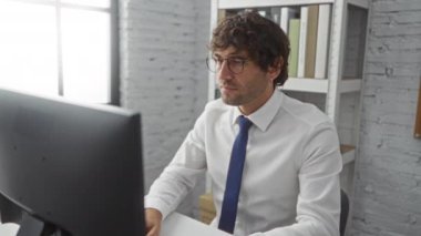 Ofisteki gözlüklü ve kravatlı genç adam, bilgisayarlı ve tabletli bir masadaki işine odaklanmış, etrafı kitaplık ve parlak bir pencereyle çevrili..