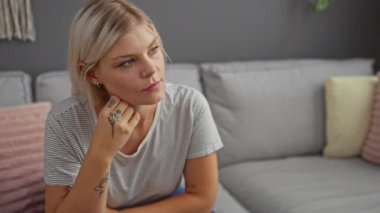 Genç bir kadın düşünceli bir şekilde oturma odasında oturuyor pastel yastıklar ve gri bir kanepeyle süslenmiş, çizgili bir gömlek giyiyor ve elinde narin dövmeler sergiliyor..