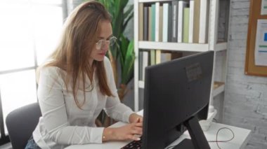 Masa başında çalışan bilgisayar, kitaplık ve geniş pencereli bir kadın odaklanmış ve profesyonel bir ev ortamı yaratıyor..