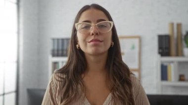 Ofis ortamında, gözlüklü, uzun kumral saçlı, pencerenin yanında duran, modern bir iş yerinde güven ve profesyonellik taşıyan bir kadın..