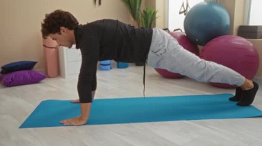 Spor salonunun içinde yoga minderinde egzersiz yapan, bilek ağrısı çeken, fitness topları ve yoga aksesuarlarıyla çevrili, odaklanmış bir çalışma ortamını taşıyan genç bir adam..