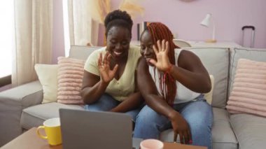 İki gülümseyen Afrikalı Amerikalı kadın, Pastel dekorlu rahat bir oturma odasında, kardeşlik ve dostluk bağlarını güçlendiren bir video görüşmesinin keyfini çıkarıyorlar..