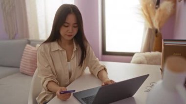 İnternetten alışveriş yapan bir kadın, yumuşak pastel dekorlu rahat bir evde kredi kartı ve laptop kullanıyor.