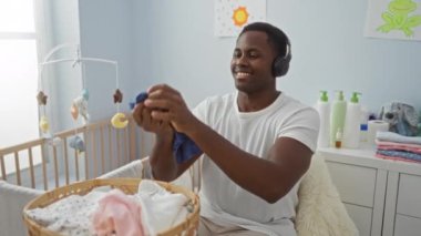 Kulaklık takan, bebek kıyafetlerini yatak odasında katlayan, oyuncaklar ve bebek eşyalarıyla çevrili, sıcak ve özenli genç bir adam..
