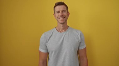 Parlak sarı bir arkaplanda duran adam, rahat gri bir tişörtle güvenle gülümsüyor, çekicilik ve pozitif tavır sergiliyor..