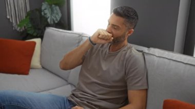 Oturma odasında el ele otururken rahat hissetmeyen bir adam. Günlük giysiler içinde. Tarafsız dekorasyon, modern mobilyalar görünür, sakin atmosfer.