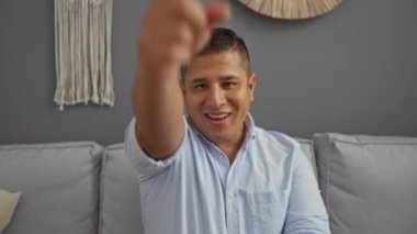 Genç adam kameraya bakıp gülümsüyor ve şık dekore edilmiş bir oturma odasında koltukta oturuyor..