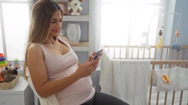 Bebek odasındaki rahat evde, yatakta oturan hamile bir kadın. Etrafı oyuncaklarla çevrili. Pencereden yansıyan parlak doğal ışık.