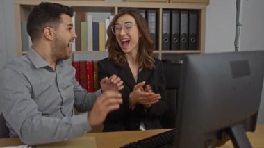 Erkek ve kadın ofis çalışma alanındaki bilgisayara el çırpıyor ve gülümsüyor, kitaplar ve ciltlerle çevrili, başarıyı coşku ve takım çalışmasıyla kutluyorlar.