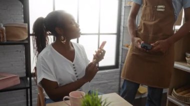 Önlüklü adama gülümseyen akıllı telefonlu bir kadın kapalı kafede elinde kart okuyucusuyla doğal ışık ile gülümsüyor.