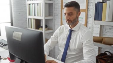 Beyaz gömlek ve mavi kravat giyen, raflar ve kitaplarla çevrili, modern çalışma alanındaki bilgisayar ekranına odaklanmış bir adam..