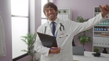 Genç İspanyol erkek doktor, modern bir klinikte güvenle gülümsüyor, elinde bir pano, parmakları havada, kapalı alanda bitkiler ve tıbbi ekipmanlarla..