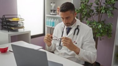 Gözlüklü doktor hastane ofisinde dizüstü bilgisayarda çalışır. Etrafı bitkiler ve tıbbi malzemelerle çevrilidir..