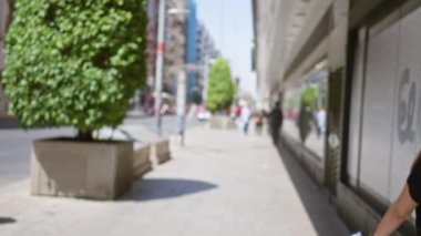 Bulanık yayalar açık şehir kaldırımlarında yürüyorlar. Arka planda yeşil alan ve binalar var.