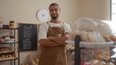 Fırın odasında sakallı genç adam çapraz kollarla gülümsüyor ve el hareketi yapıyor. Etrafı ekmek ve hamur işleriyle çevrili, fırının arka planında kahverengi bir önlük giyiyor.