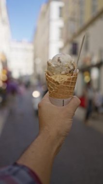 Bulanık kentsel sokak ortamında elinde dondurma külahı tutan bir adam, arka planda insanlar, sıradan bir açık hava anını vurguluyor..