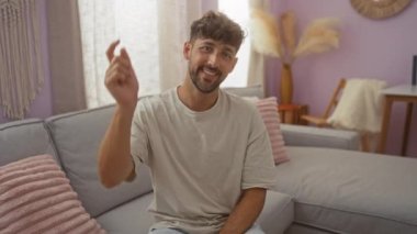 Sakallı, kısa saçlı, gülümseyen ve kameraya bakan genç bir adam açık mor duvarlı, minimalist dekorlu, rahat bir oturma odasında gri bir kanepede oturuyor.