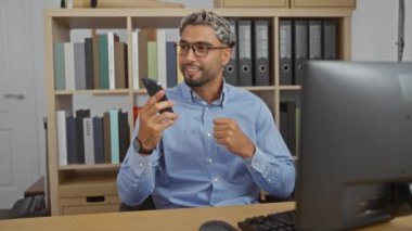 Ofisteki yakışıklı genç arka planda kitaplık ve masaüstü bilgisayarıyla akıllı telefondan konuşuyor.