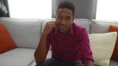 Pembe gömlekli genç adam, modern dekorlu rahat bir oturma odasında kanepede otururken gülümsüyor ve işaret ediyor..