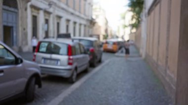Roma, İtalya 'daki kaldırım taşı sokağının bulanık manzarası, park halindeki arabaların ve belli belirsiz yayaların yer aldığı, tarihi bir şehir ortamındaki bokeh etkisini vurguluyor..