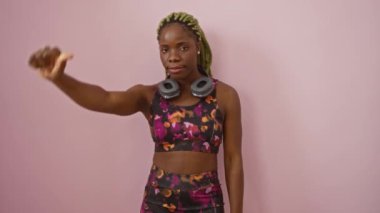 Spor kıyafetli genç Afro-Amerikan kadın kulaklıklarıyla olumsuz bir hoşnutsuzluk ve reddedilme belirtisi sergiliyor. Tecrit edilmiş pembe arka plana kızgın bakıyor.