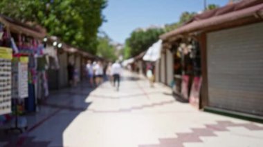 Güneşli bir pazar caddesinde arka planda bulanık dükkanlar ve ağaçlarla yürüyen insanlar, hareketli bir tatil ortamı yaratıyor.