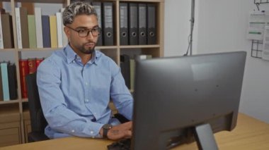 Ofiste çalışan genç bir adam, yorgunluktan gözlerini ovuşturuyor, iş yerindeki bir masada arkasında kitap ve ciltlerle dolu bir bilgisayarla oturuyor.