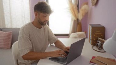 Evde dizüstü bilgisayar kullanan genç adam bilgisayarı kapatıyor ve temiz bir masanın üzerindeki kitaplarla çevrili, kapalı bir oturma odasına uzanıyor..