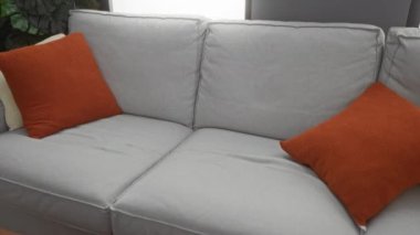 Genç adam, modern oturma odasındaki turuncu yastıklı rahat bir kanepede sıkıntılı bir şekilde oturuyor..