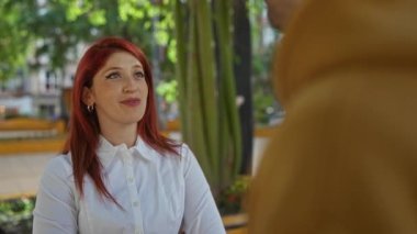 Güneşli açık hava parkında, canlı yeşil ağaçlarla gülümseyen, beyaz tişört giyen, sarılı insanlarla sohbet eden, neşeli ve rahat bir atmosfer yaratan bir kadın..