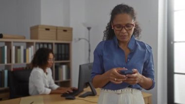 Akıllı telefon kullanan bir kadın ve arka planda klasörler ve depolama kutularıyla ofiste çalışan başka bir kadın.