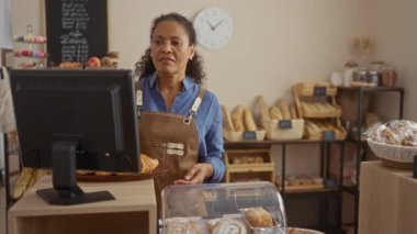 Pastanede çalışan bir kadın, müşterilere taze hamur işleri sunan, kasanın yanında duran ekmek ve çörekler kapalı bir ortamda raflarda sergileniyor..