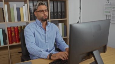 Ofis odasında sakallı genç bir adam bilgisayarla masaya uzanıyor ve gözlüklerini çıkarıyor. Arka planda kitaplıklarla bitkin görünüyor.