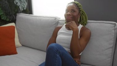 Evdeki gri kanepede oturan, turuncu yastıklı, kot pantolon giyen, beyaz bluzlu, bitki süsleme odası dekorasyonu yapan bir kadın.