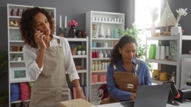 Ev dekorasyon dükkanında çalışan kadınlar, biri telefonla konuşuyor ve diğeri dizüstü bilgisayar kullanıyor, çeşitli dekoratif eşyalar ve bitkilerle çevrili.