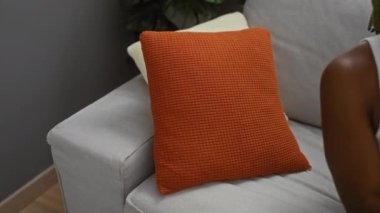 Kanepede, rahat bir dairede, turuncu yastıkla rahat bir şekilde uzanan, sıradan bir bluz giyen ve örgü ören bir kadın..