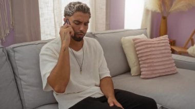 Sakallı genç adam bir telefon görüşmesini bitirdikten sonra hayal kırıklığına uğramış görünüyor. Modern bir oturma odasında, yumuşak pastel renklerle süslenmiş rahat bir koltukta oturuyor.