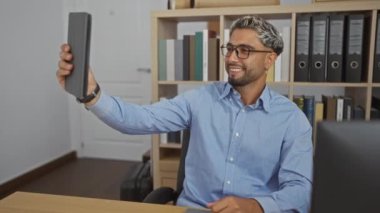 Genç Arap işyerindeki tablette selfie çeken, gözlüklü ve mavi gömlekli, gülümseyen, masasında oturan, kapalı alandaki raflarda kitap ve klasörlerle çevrili genç bir adam..