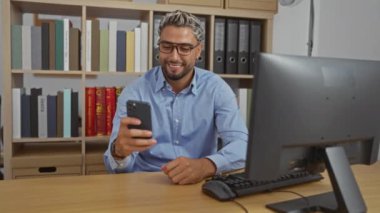 Sakallı ve gözlüklü genç adam modern ofisteki akıllı telefondan bir video görüşmesi yapıyor. Arka planda kitaplıklar var, gülümsüyor ve sohbete dalmış.