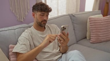 Genç adam modern bir oturma odasında rahat bir kanepede dinlenirken akıllı telefon kullanıyor.