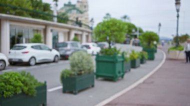Monako caddesinin bulanık görüntüsü lüks arabaları, yeşillikleri ve yayaları çekiyor. Odaklanmamış, bokeh dolu bir açık hava ortamında şehir zarafetini sergiliyor..
