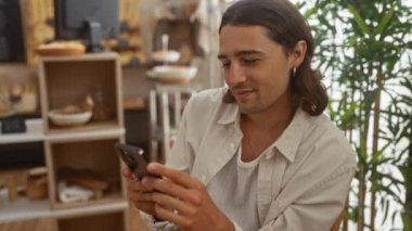 Uzun saçlı genç adam fırında akıllı telefon kullanıyor. Arkasında ekmek rafları ve hamur işleri var.