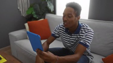 Video görüşmesi sırasında rahat oturma odasında tablet kullanan, çizgili gömlek giyen ve gri bir koltukta oturan, dekoratif bitkiler ve turuncu minderlerle ev ortamını geliştiren genç bir adam..