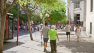 Güneşli bir caddede yürüyen, erkek, kadın, ağaç, bina ve arka planda bir otobüsün yer aldığı bulanık bir açık hava sahnesi.