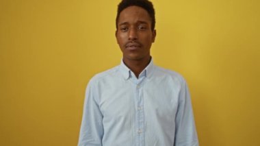 Afro-Amerikalı genç bir adam gömleğini giyerek mutlu bir gülümseme ve izole edilmiş sarı arka planda dostça bir selamlama ile selam veriyor.