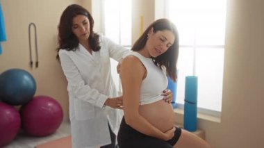 Hamile bir kadın spor salonunda arka planda egzersiz topları ve yoga minderleriyle kadın terapi görüyor. Bu da destekleyici bir sağlık ve spor ortamı olduğunu gösteriyor.